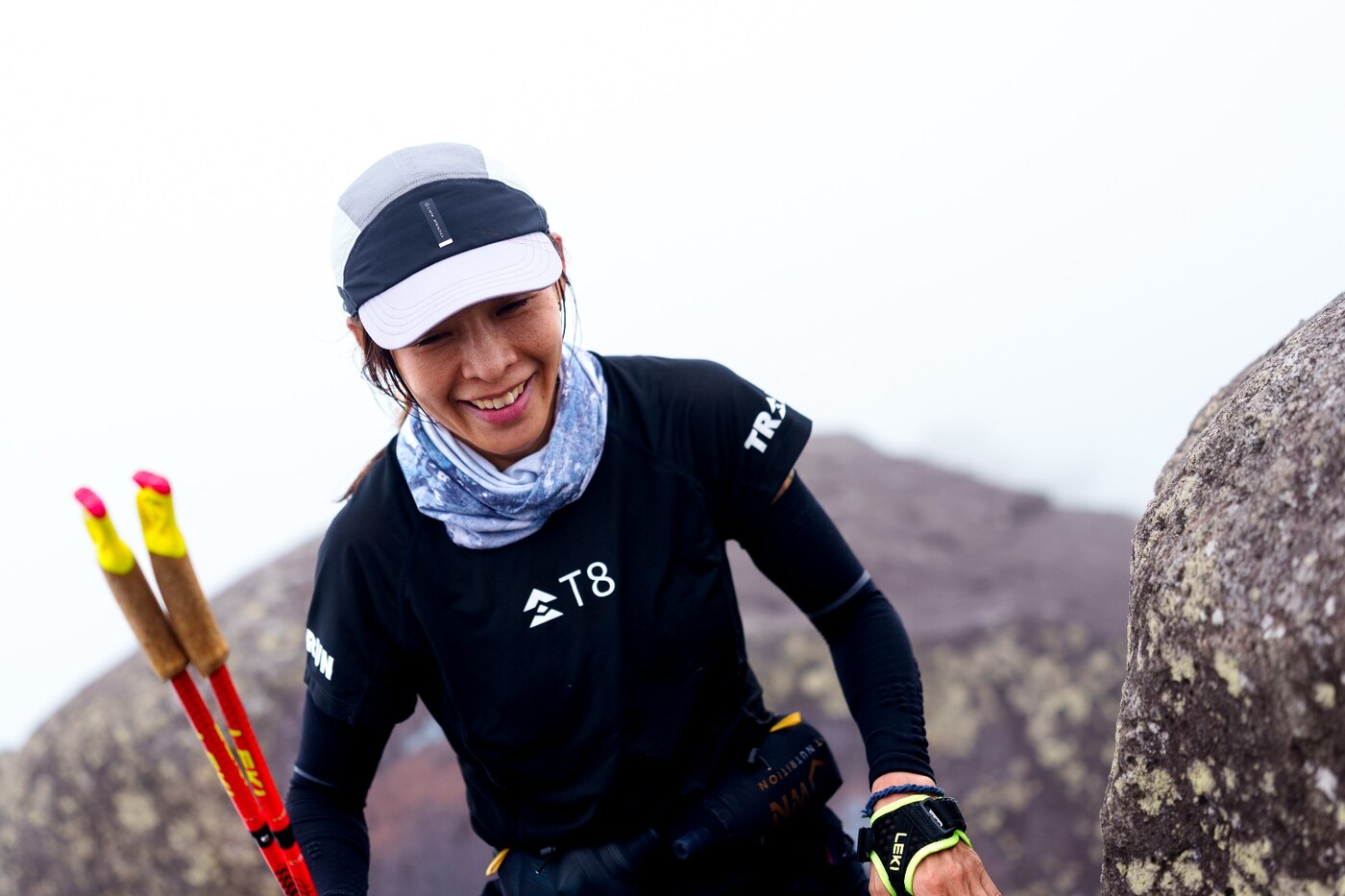 Smiling woman runner with poles climbing a misty rocky summit